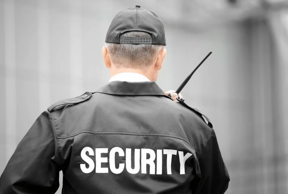 A security guard in a black uniform and cap stands with his back to the camera, holding a radio at an outdoor location.