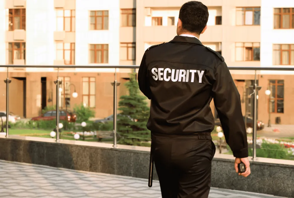 A security guard in a black uniform walks away, patrolling a modern building's entrance with greenery and cars in the background.