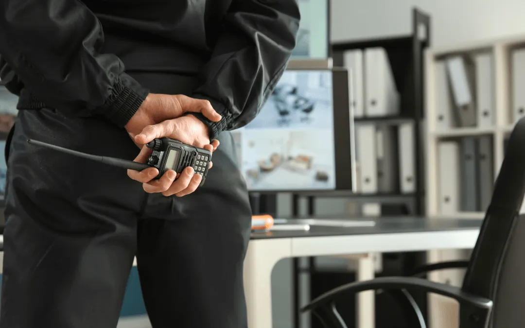 A person in a black outfit holding a radio, standing with their back to the camera in an office setting with monitors and shelves in the background.