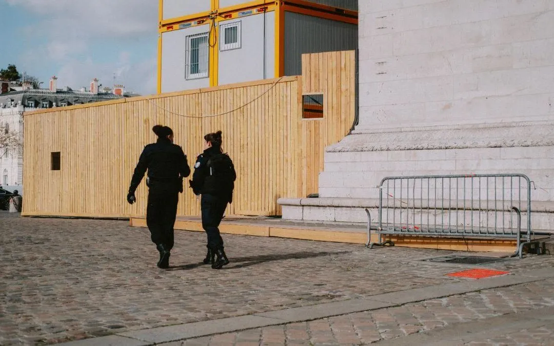 Two police officers walking on a cobblestone path near a wooden barrier and a modern structure.